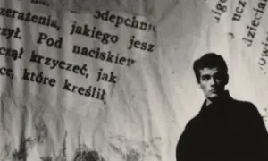Młody mężczyzna, być może Hanuszkiewicz, stoi na tle podartych kartek z tekstem.