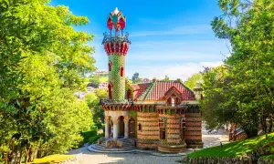 Kolorowy budynek z wieżą, otoczony zielenią. Ciekawostki o Antonio Gaudí często dotyczą jego unikalnych projektów, jak ten.