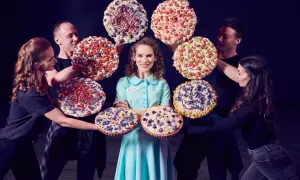 Jenna z musicalu "Waitress" otoczona przez aktorów z kolorowymi, udekorowanymi ciastami.