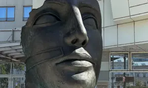 Brązowa, fragmentaryczna rzeźba twarzy Igora Mitoraja na tle nowoczesnego budynku.