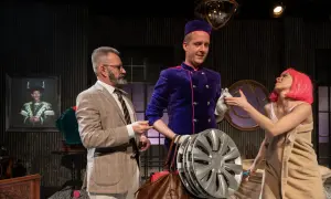 Grzegorz Wiśniewski reżyser, w teatralnej scenografii, z aktorką w różowej peruce i aktorem w garniturze.