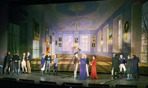 Scena salon warszawski. Grupa postaci w strojach z epoki zgromadzona w eleganckim wnętrzu z fortepianem i obrazami.