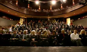 Widownia teatru wypełniona po brzegi. Ludzie siedzą w rzędach, czekając na spektakl. Widać kilka osób na pierwszym planie, a za nimi tłum.