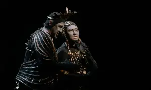 Dwaj aktorzy w teatralnych strojach, przypominających kostiumy z opery Carmen, z pomalowanymi na szaro twarzami, w mroku sceny.