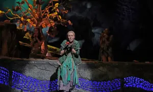 Aktorka w zielonej sukni trzyma jabłko, symbol grzechu w operze "Raj utracony". W tle drzewo i inne postacie.