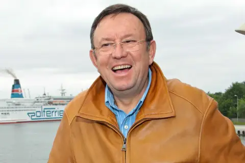 Uśmiechnięty Zbigniew Buczkowski w brązowej kurtce, na tle promu Polferries. Jego filmy, seriale i programy to klasyka polskiej rozrywki.