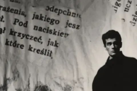 Młody mężczyzna, być może Hanuszkiewicz, stoi na tle podartych kartek z tekstem.