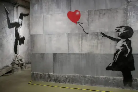 Dzieło Banksy'ego: dziewczynka sięga po czerwone serce, obok gimnastyczka na rękach. Kim jest Banksy? Tajemniczy artysta.
