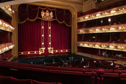 Wnętrze teatru z czerwonymi aksamitnymi zasłonami i lożami. Słychać śpiewaka operowego, który przygotowuje się do występu.