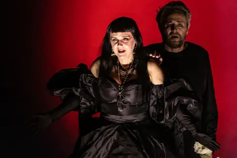 Dramatyczna scena z opery Karola Szymanowskiego. Kobieta w czarnej sukni i mężczyzna z wyrazem przerażenia na twarzy.