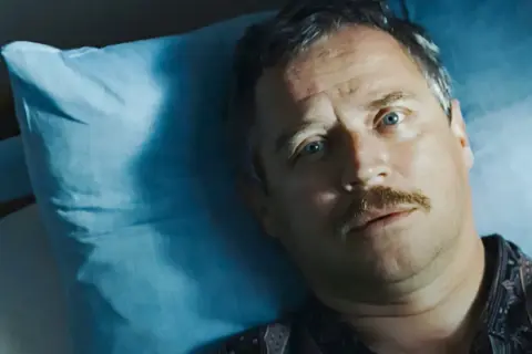 Mężczyzna z wąsami leży na niebieskiej poduszce, jego oczy patrzą w górę. Może to scena z filmu "Dzień świra", gdzie reżyser ukazuje codzienne zmagania bohatera.