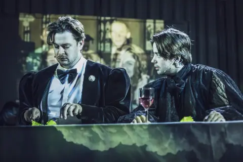 Dwóch mężczyzn przy stole, jeden w czarnym aksamicie, drugi w koronkowej szacie. Może to Hamlet i Ofelia?