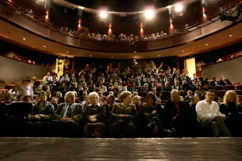 Widownia teatru wypełniona po brzegi. Ludzie siedzą w rzędach, czekając na spektakl. Widać kilka osób na pierwszym planie, a za nimi tłum.