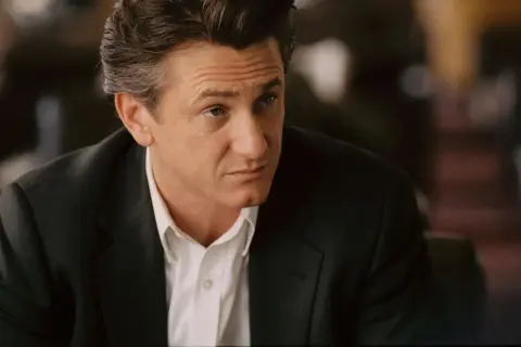 Sean Penn w czarnym garniturze i białej koszuli, zamyślony. Jego role w filmach zapadają w pamięć.