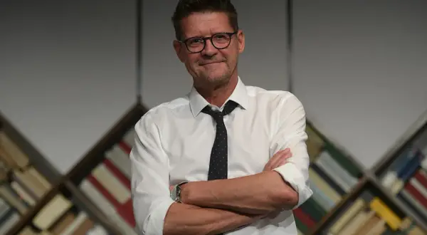 Wojciech Malajkat w białej koszuli i krawacie, z okularami na nosie, stoi na tle regałów z książkami. Jego filmy to klasyka polskiego kina.