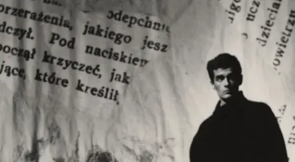 Młody mężczyzna, być może Hanuszkiewicz, stoi na tle podartych kartek z tekstem.