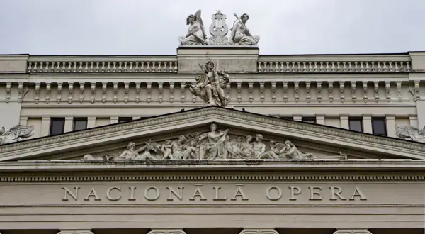 Elegancka fasada budynku opery w stylu neoklasycyzmu, ozdobiona rzeźbami i napisem "NACIONĀLĀ OPERA".