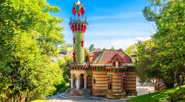 Kolorowy budynek z wieżą, otoczony zielenią. Ciekawostki o Antonio Gaudí często dotyczą jego unikalnych projektów, jak ten.