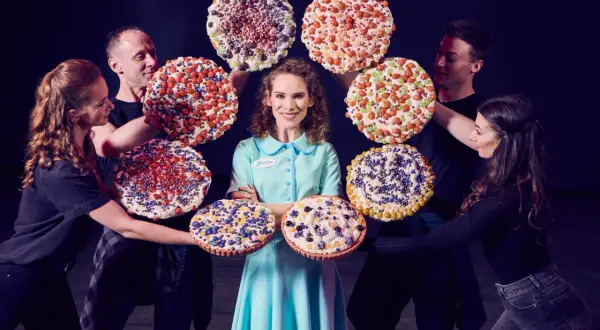 Jenna z musicalu "Waitress" otoczona przez aktorów z kolorowymi, udekorowanymi ciastami.