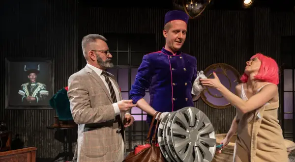 Grzegorz Wiśniewski reżyser, w teatralnej scenografii, z aktorką w różowej peruce i aktorem w garniturze.