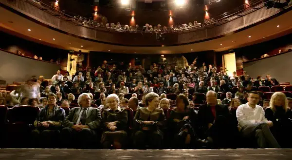 Widownia teatru wypełniona po brzegi. Ludzie siedzą w rzędach, czekając na spektakl. Widać kilka osób na pierwszym planie, a za nimi tłum.