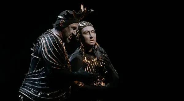 Dwaj aktorzy w teatralnych strojach, przypominających kostiumy z opery Carmen, z pomalowanymi na szaro twarzami, w mroku sceny.