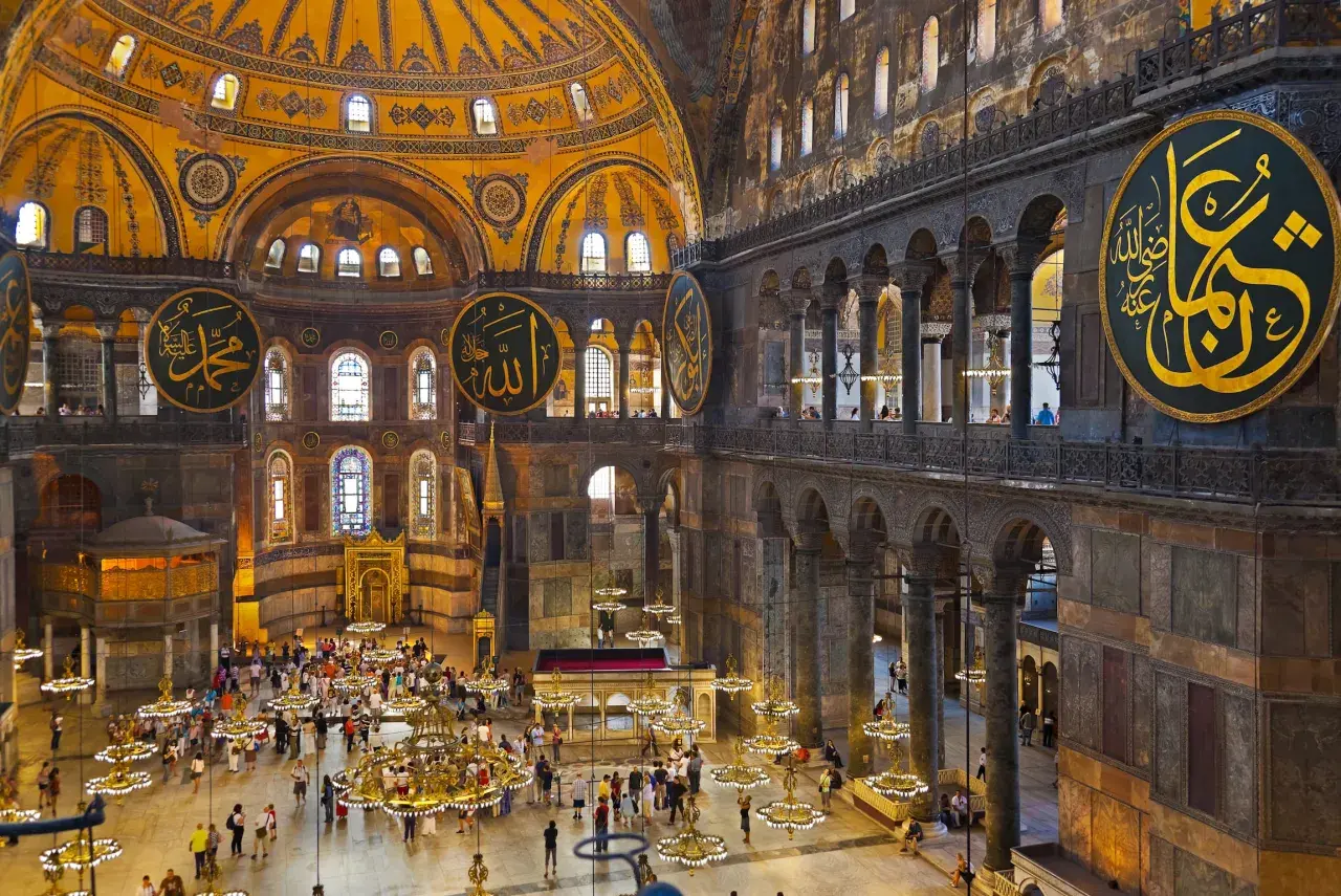 Wnętrze Hagia Sophia z widokiem na kopułę, kaligrafię i turystów. Plan budowli ukazuje jej monumentalność.