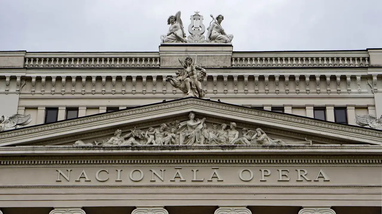 Elegancka fasada budynku opery w stylu neoklasycyzmu, ozdobiona rzeźbami i napisem "NACIONĀLĀ OPERA".