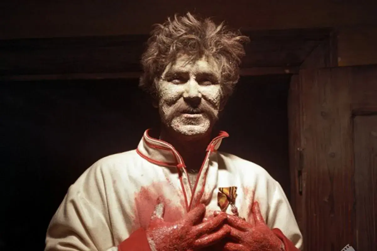 Mężczyzna z twarzą pokrytą białą substancją, w białej kurtce z czerwonymi zdobieniami i zakrwawionymi rękami, trzyma medal. Może to być symboliczny czepiec wesele.