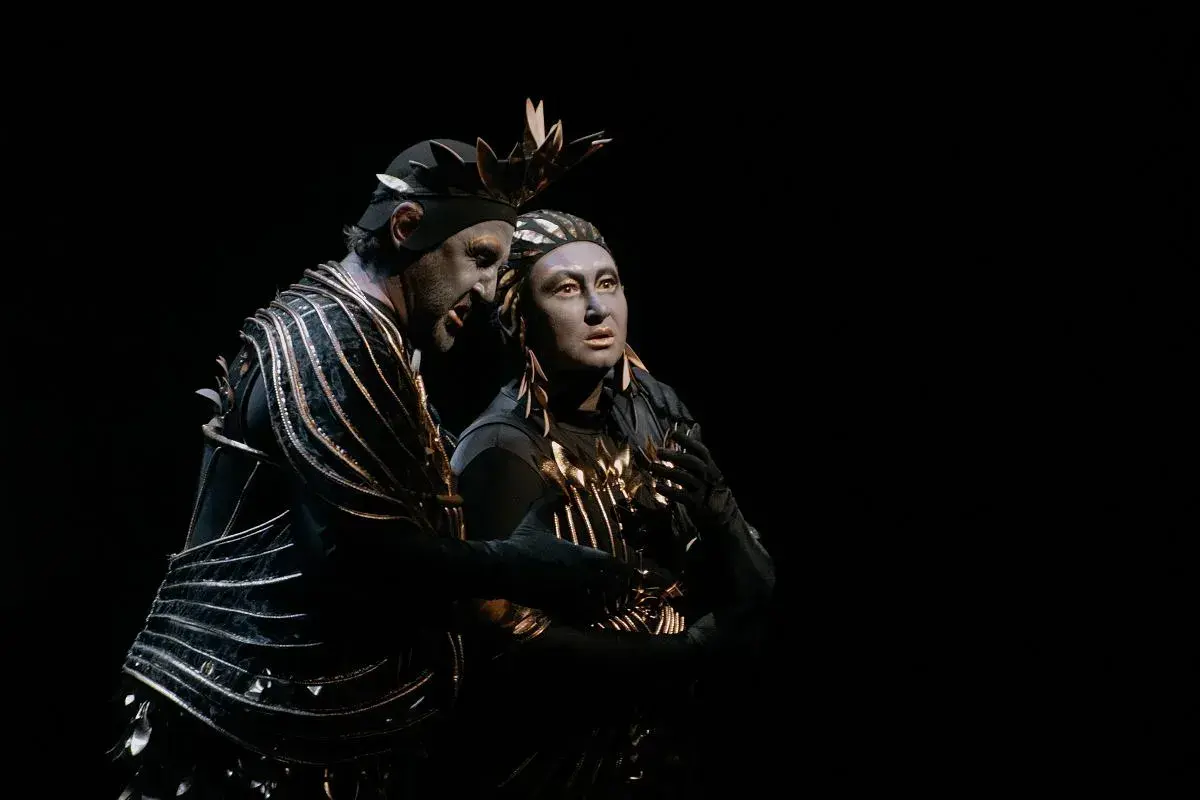 Dwaj aktorzy w teatralnych strojach, przypominających kostiumy z opery Carmen, z pomalowanymi na szaro twarzami, w mroku sceny.