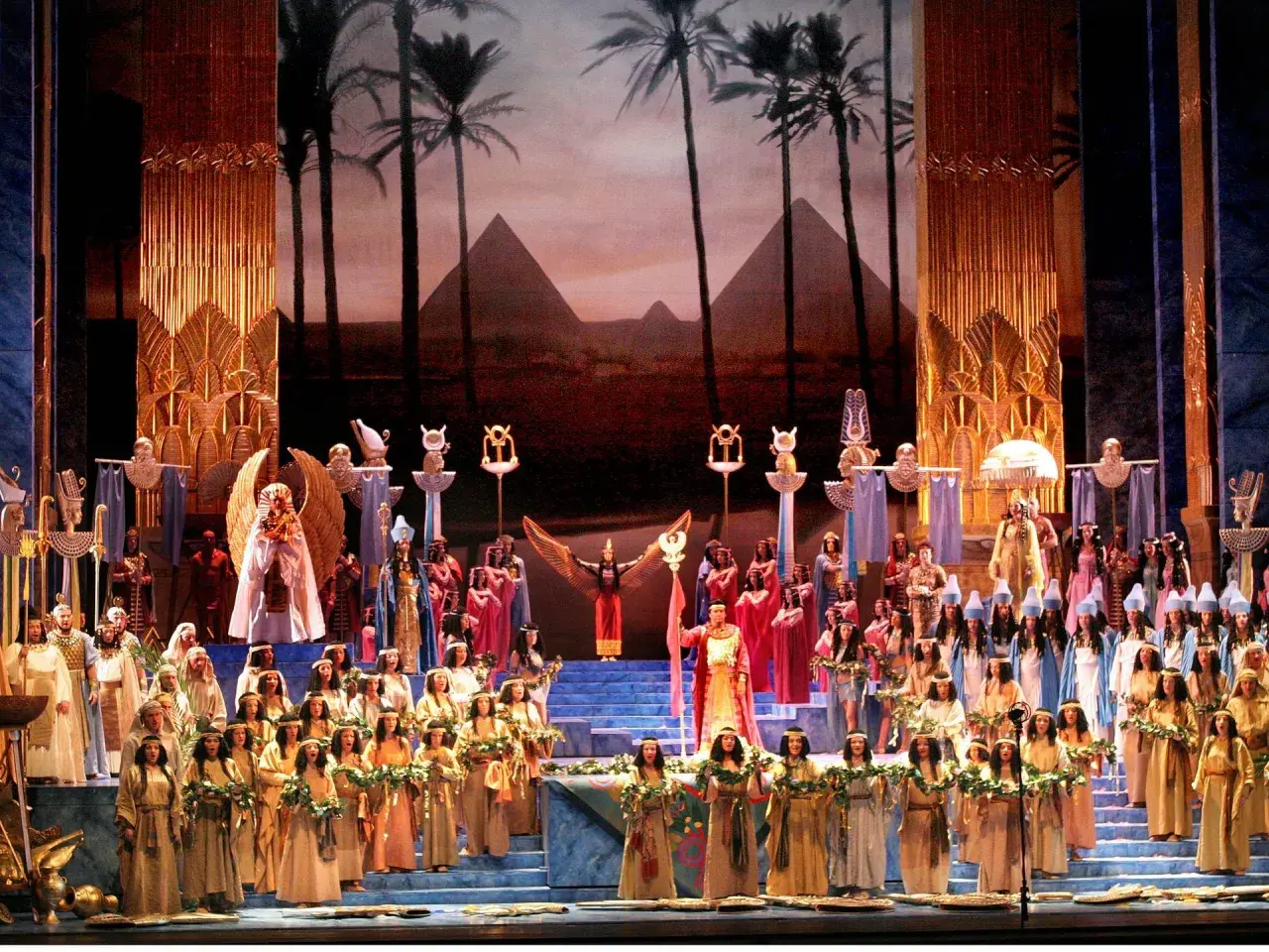 Scena z opery Aida: tłum w starożytnych strojach, piramidy i palmy w tle.