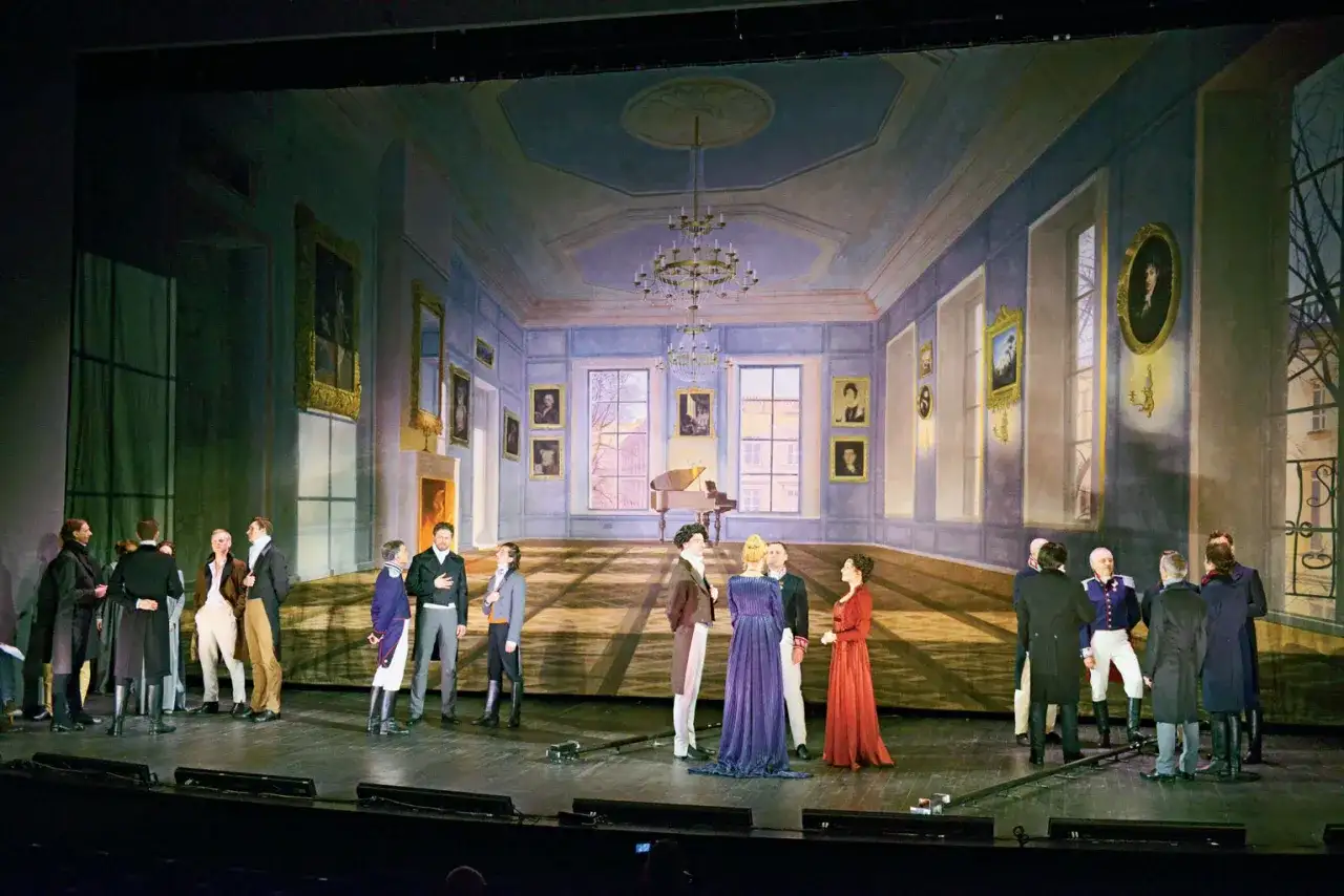 Scena salon warszawski. Grupa postaci w strojach z epoki zgromadzona w eleganckim wnętrzu z fortepianem i obrazami.