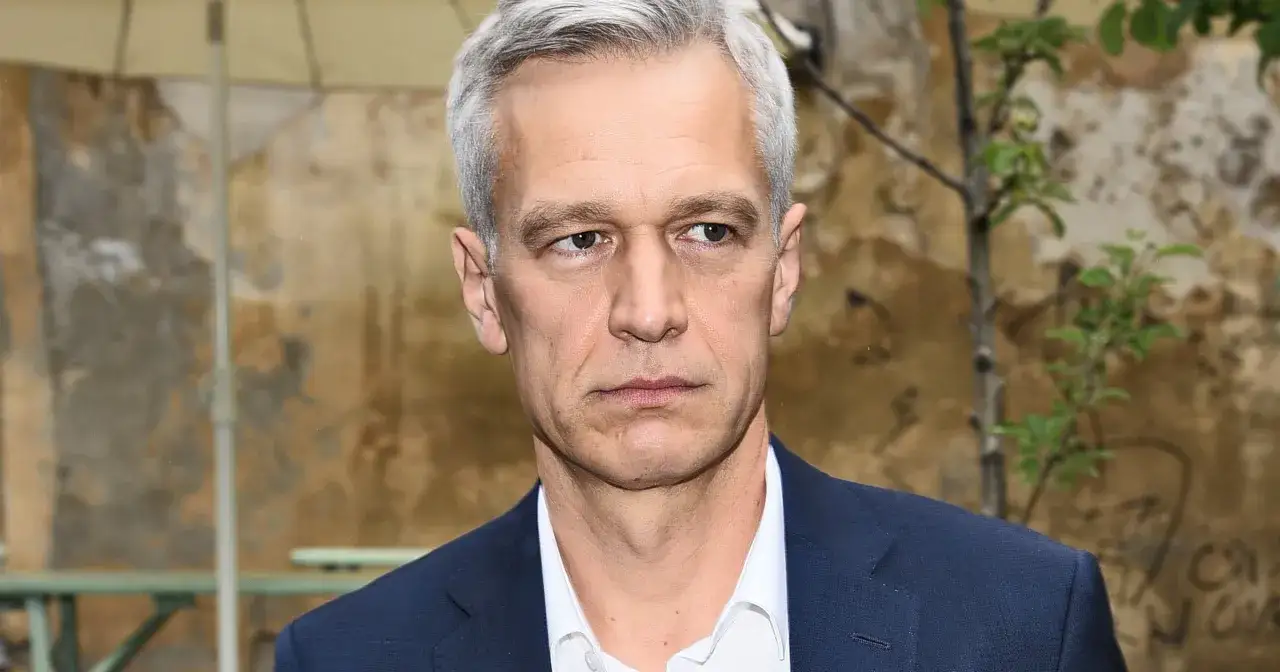 Michał Żebrowski w garniturze, znany z wielu filmów, spogląda w bok na tle starej ściany i drzewa.