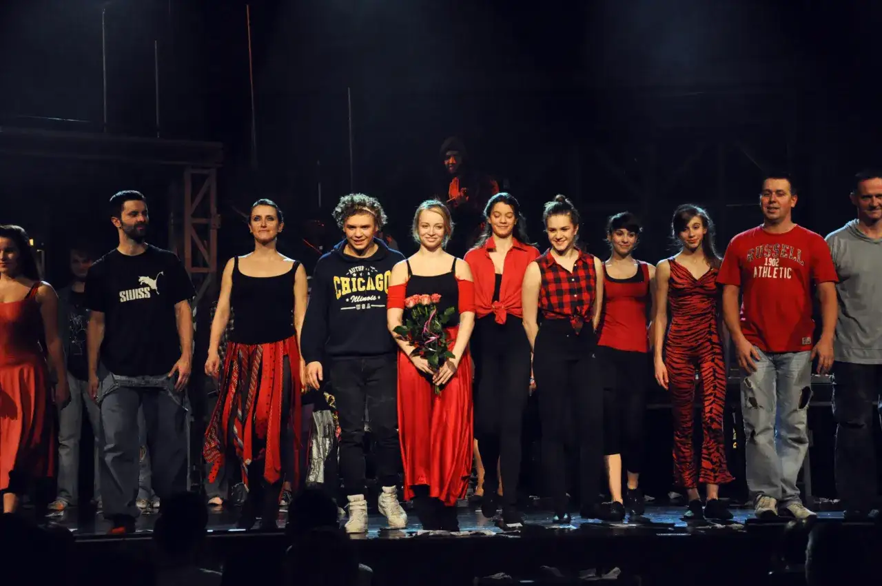 Aktorzy teatru buffo na scenie, w kolorowych strojach, pozdrawiają publiczność.