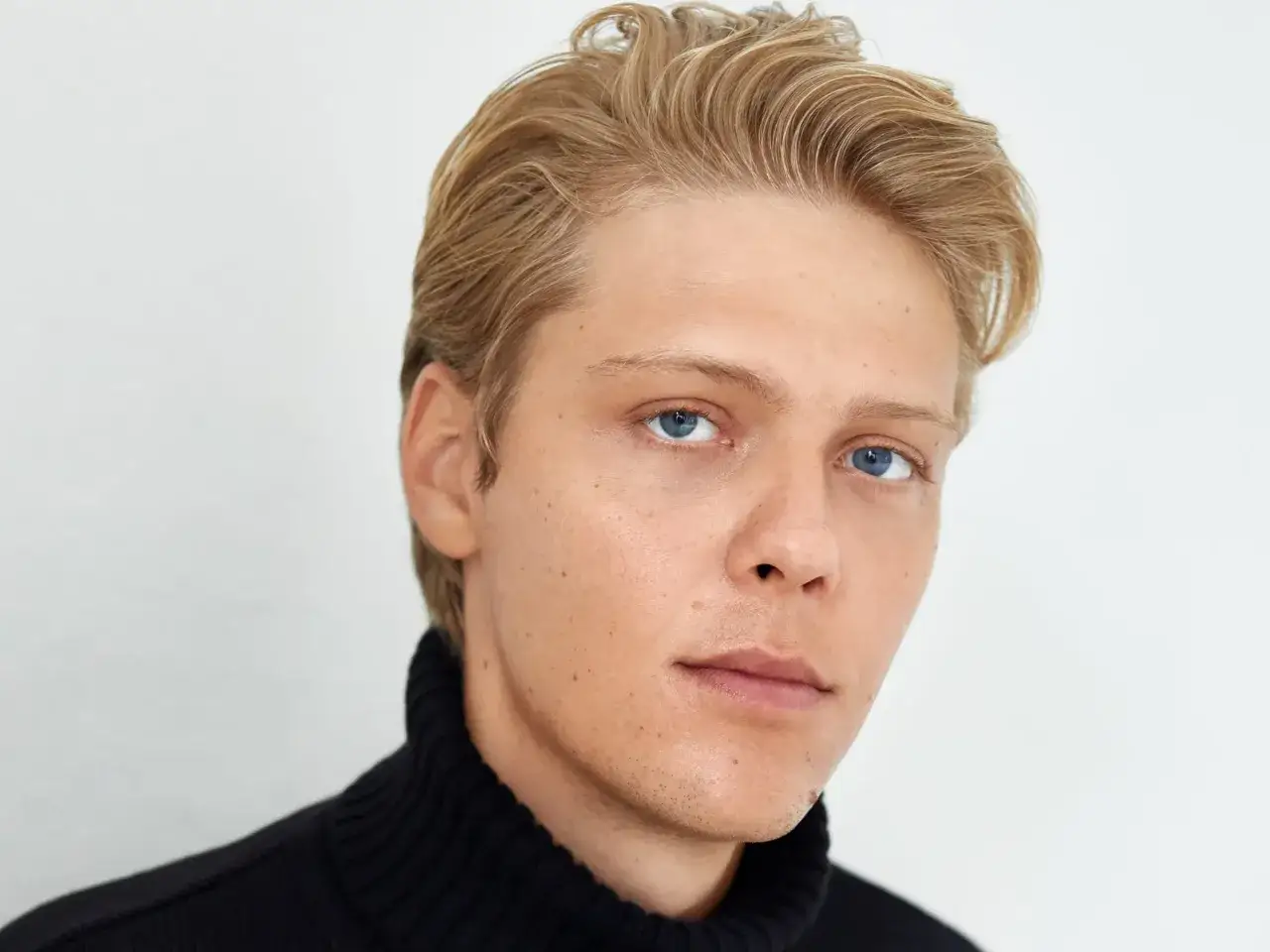 Młody mężczyzna o blond włosach i niebieskich oczach, ubrany w czarny golf. Może to być Jan Holoubek, znany z filmów, seriali i programów.
