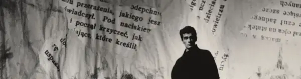 Młody mężczyzna, być może Hanuszkiewicz, stoi na tle podartych kartek z tekstem.