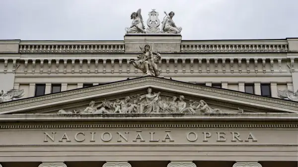 Elegancka fasada budynku opery w stylu neoklasycyzmu, ozdobiona rzeźbami i napisem "NACIONĀLĀ OPERA".
