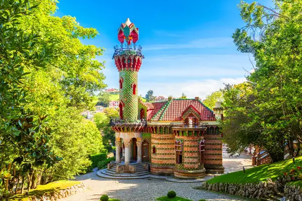 Kolorowy budynek z wieżą, otoczony zielenią. Ciekawostki o Antonio Gaudí często dotyczą jego unikalnych projektów, jak ten.