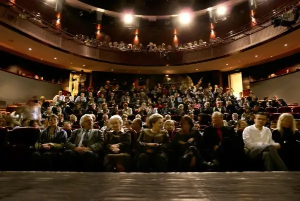 Widownia teatru wypełniona po brzegi. Ludzie siedzą w rzędach, czekając na spektakl. Widać kilka osób na pierwszym planie, a za nimi tłum.